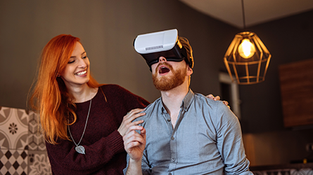 homem ruivo de camisa social inza utilizando um óculos de realidade virtual aumentada ao lado de uma mulher ruiva de blusa marrom que segura em seus ombros. O ambiente é parecido ai de uma sala de casa