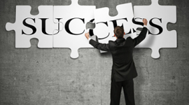 Foto de um homem de terno que está de costas. Ele monta um quebra-cabeça na parede com a palavra success. Ele está colocando a última peça com as letras CE da palavra success.