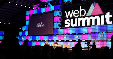 Foto. Duas pessoas sentadas em cadeiras no palco do web summit. Atrás delas um telão e uma parede com as cores azuis e lilás e um letreiro com o nome do evento. A platéia está na frente dos palestrantes. 