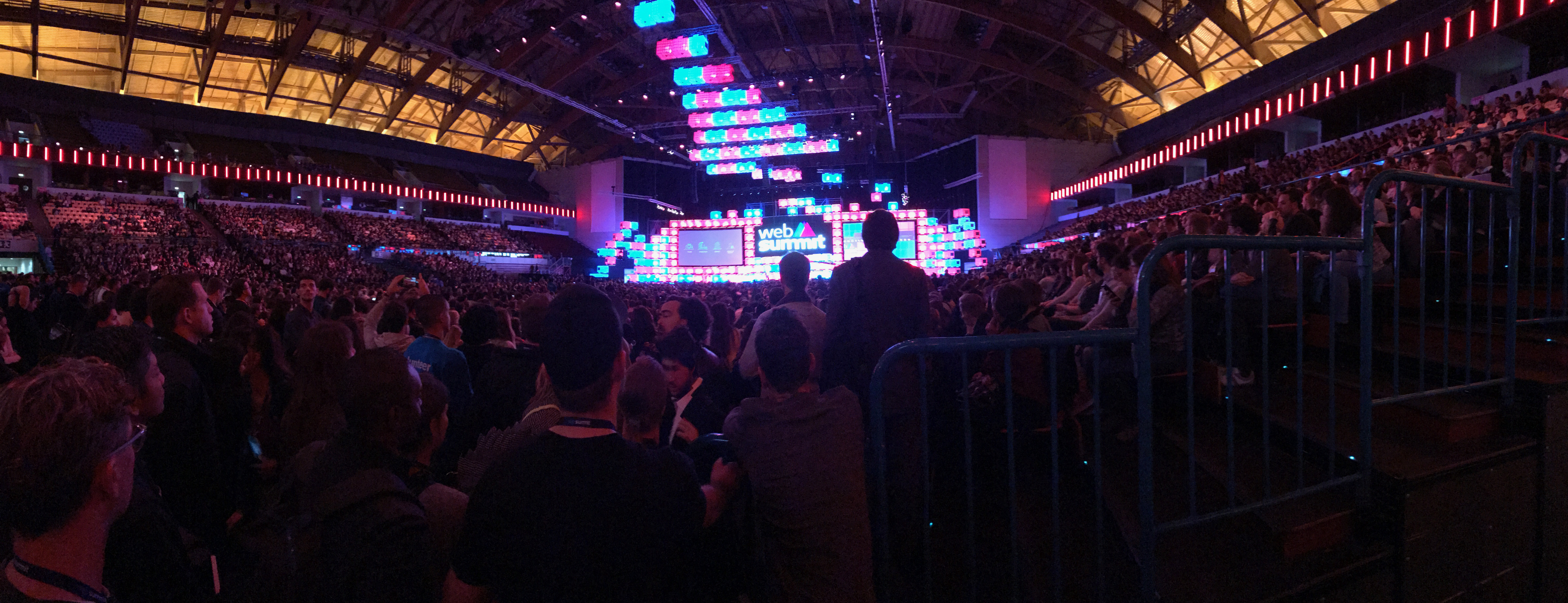 Foto de uma arena, repleta de pessoas sentadas e em pé. Ao fundo, as luzes de palco em rosa e azul. 