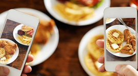 Foto. Três pratos de comida brancos e segundo plano. Duas pessoas seu segurando seus celulares e na tela dos celulares aparece o prato de comida mais nítido.