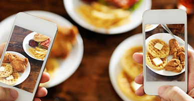 Foto. Três pratos de comida brancos e segundo plano. Duas pessoas seu segurando seus celulares e na tela dos celulares aparece o prato de comida mais nítido. 