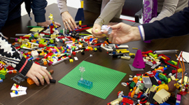Foto. Mãos de pessoas sob uma mesa segurando peças de lego. Em cima da mesa várias peças de lego espalhadas.