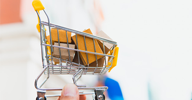 Imagem: Mão segurando uma miniatura de carrinho de compras com caixinhas. Consumidores.