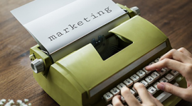 Imagem: mão escrevendo em máquina de escrever, no papel a palavra "marketing". ferramentas de marketing.