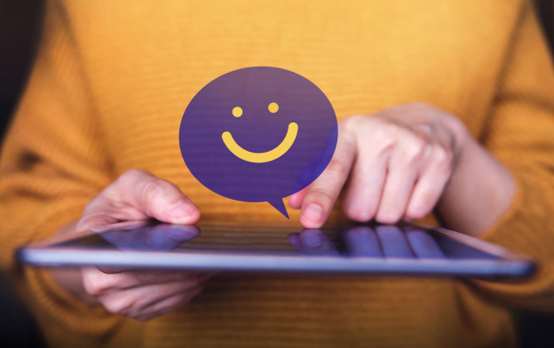 Imagem: pessoa segurando um tablet, com um balão roxo e uma carinha feliz, representando uma boa experiência digital.